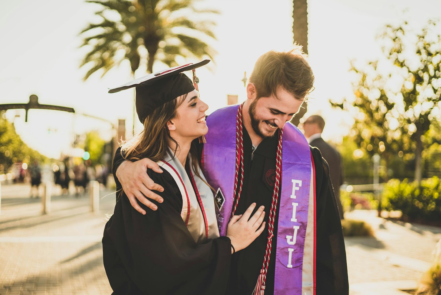 Two graduates embrace joyfully outdoors near palm trees.
